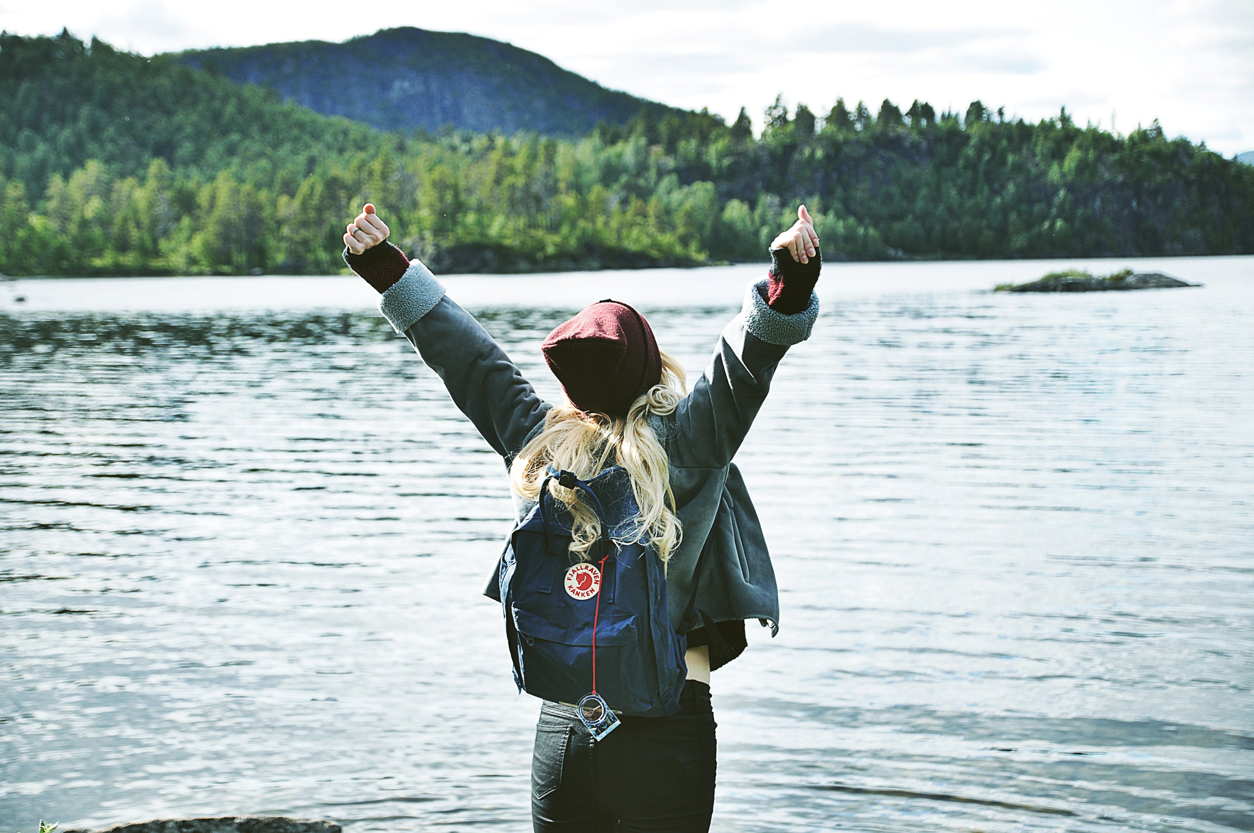 Woman in front of a large lake, with an arctic fox backpack on her back, stretching up her arms