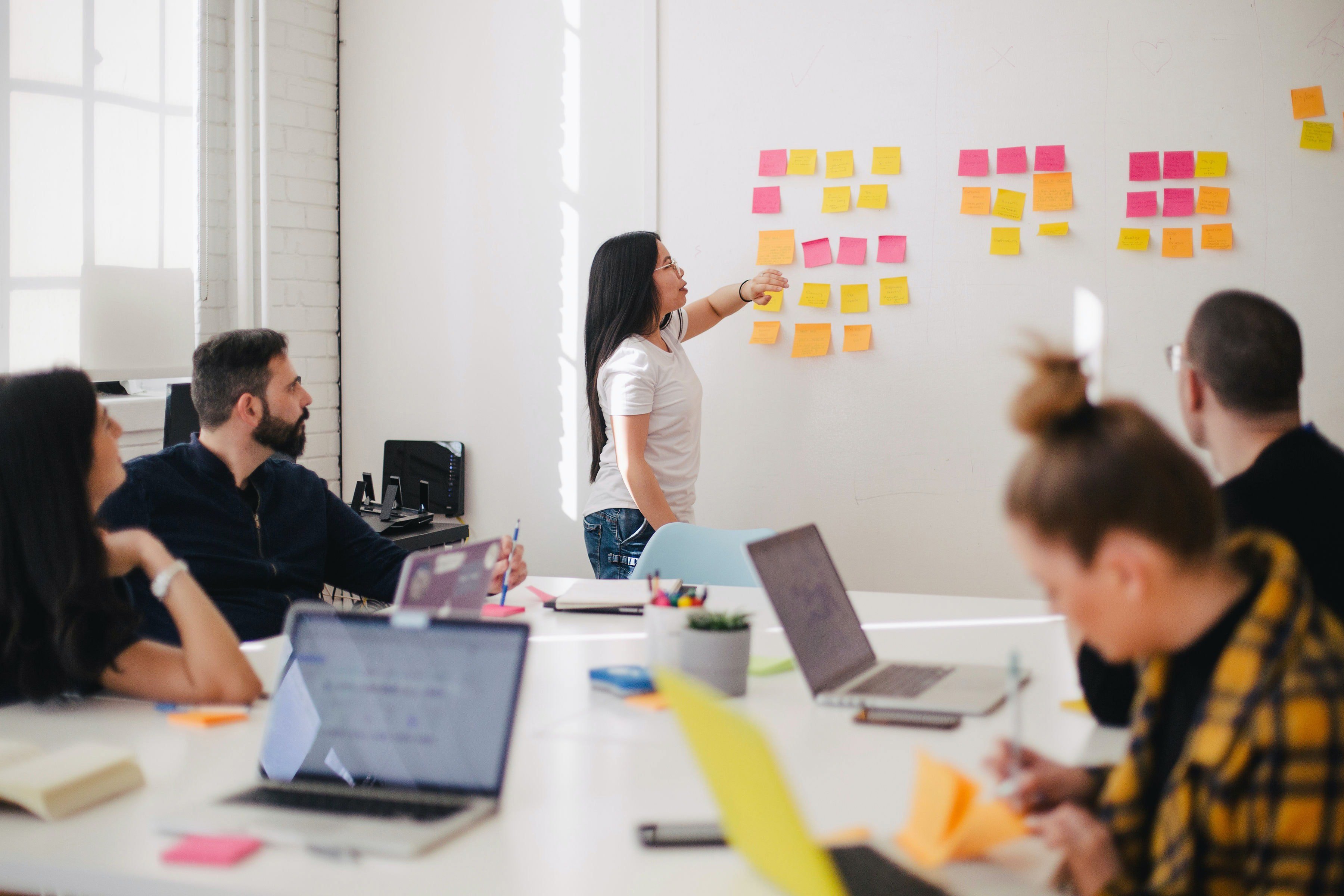 Kvinna vid whiteboard täckt i postitlappar presenterar för en grupp personer 